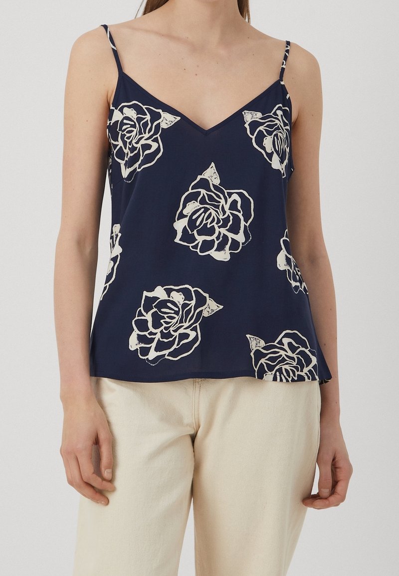 Femme portant un haut bleu marine sans manches à col en V avec de grands imprimés floraux blancs et un pantalon beige clair.