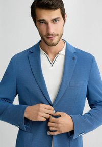 Blauer strukturierten Blazer mit Revers, einreihigem Knopfverschluss und einer Brusttasche, getragen über einem weißen Hemd mit Kragen und schwarzen Streifen.
