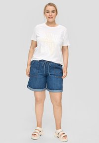 Witte t-shirt met daarop "SAND OCEAN SALT WIND" gedrukt in pastelkleuren, gecombineerd met blauwe denim shorts en beige sandalen. Casual stijl, relaxte pasvorm.