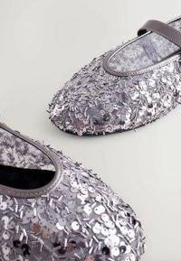 Silberne Ballettschuhe aus Pailletten mit einem strukturierten Obermaterial, runder Zehenpartie und grauem Riemen. Mit glänzenden, scheibenförmigen Verzierungen.