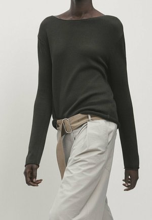 Personne portant un pull vert foncé à manches longues, rentré dans un pantalon beige avec une ceinture en tissu assortie et une boucle métallique.