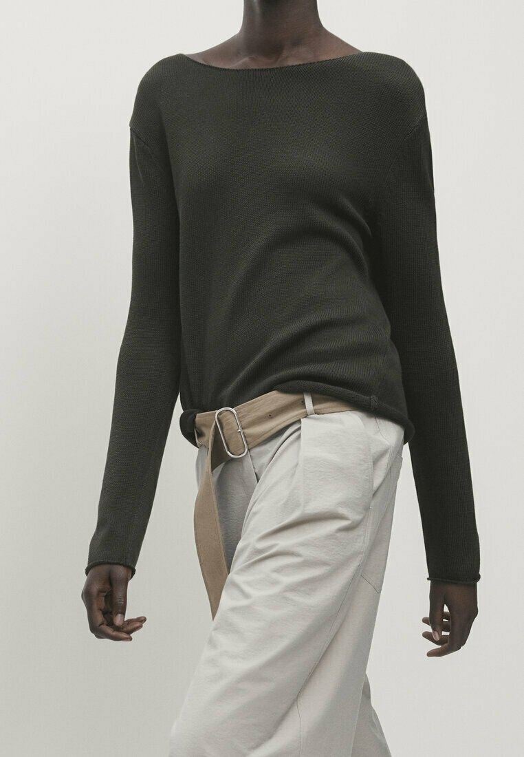 Personne portant un pull vert foncé à manches longues, rentré dans un pantalon beige avec une ceinture en tissu assortie et une boucle métallique.