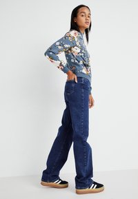 Blomstrete mønster, blå langermet topp med ribbede mansjetter og liv, kombinert med høyhårede, mørkeblå denim jeans og svarte joggesko.