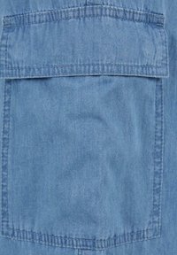 Světlomodrá džínová tkanina s velkou náprsní kapsou, šitými detaily švů a hladkou texturou.