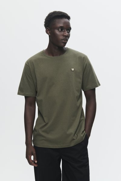 Young man wearing olive green t-shirt with small logo on chest, black pants, standing with one hand in pocket, looking to side against plain background.