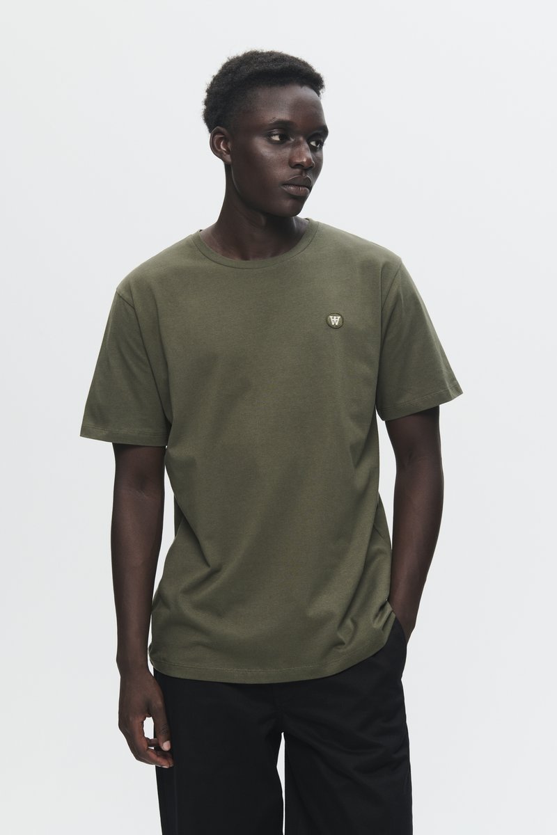 Young man wearing olive green t-shirt with small logo on chest, black pants, standing with one hand in pocket, looking to side against plain background.