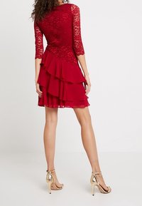 Abito rosso in pizzo con maniche a tre quarti, strati di volant all'orlo e un corpetto aderente. Abbinato a scarpe con tacco alto dorate.