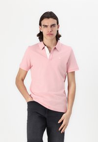 Lichtroze polo shirt met een witte kraag en een knoopsluiting met drie knopen. Korte mouwen, zachte textuur en klein geborduurd logo op de borst.