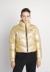 Metalická zlatá puffer bunda s vysokým l�ímcem, zipovým zapínáním a chevronovým prošíváním. Obsahuje dvě boční kapsy a elastické manžety.