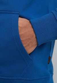 Blue sweatshirt with a kangaroo pocket, ribbed cuffs, and stitching details. Fabric appears soft with a smooth texture. Small black tag visible.