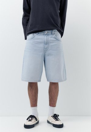 Osoba nosiaca svetlomodré denimové šortky po kolená, biele ponožky a biele tenisky s čiernymi šnúrkami na jednoduchom svetlom pozadí.