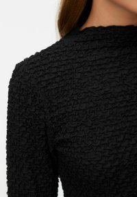 Close-up van een persoon die een zwart textuurstof top draagt met een geplooid, gerimpeld patroon, waarbij de schouder en het bovenste gedeelte van de borst te zien zijn.