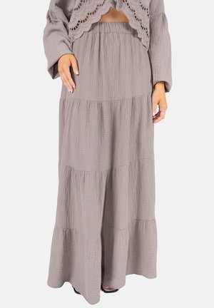 Femme portant une jupe longue à étages, texturée marron clair, associée à un haut assorti avec des détails en dentelle et des manches larges.