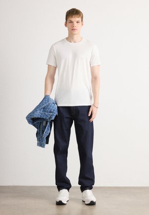T-shirt en coton blanc associé à un pantalon en denim bleu foncé. Une veste en denim bleu est tenue dans la main gauche. Des baskets blanches sont portées.