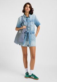 Ljusblå denimkortärmad jumpsuit med knappar framtill, fickor och bälte i midjan, tillsammans med gröna sneakers och en randig toteväska.
