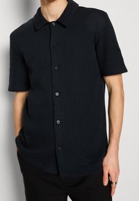 Schwarzes kurzärmliges Hemd mit gerippter Struktur, klassischem Kragen und blauen Knöpfen, das eine lockere Passform und zwei Seitentaschen aufweist.