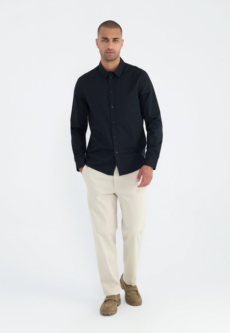 Camisa negra de manga larga con botones, pantalones beige y zapatos marrones. La camisa presenta un cuello clásico y una textura suave.
