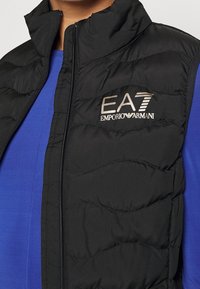 Svart pufferväst med kviltat mönster och stående krage, med en silvrig logotyp "EA7 EMPORIO ARMANI" på vänster bröst. Bärs över en blå skjorta.