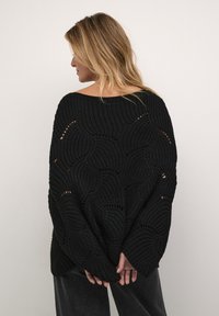 Svart stickad oversized tröja med öppna cirkulära mönster, bred halsringning och batwingärmar. Den strukturerade tyget ger en mysig känsla.