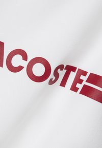 Un tessuto in cotone bianco presenta il grande testo rosso "LACOSTE" con una striscia rossa orizzontale. La superficie è liscia e le lettere hanno una leggera finitura lucida.