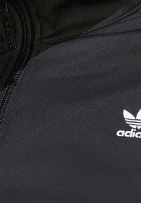 Černá bunda Adidas s bílým logo trefoil na hrudi a viditelným černým zipem na levé straně.