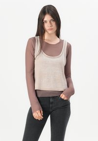 Vaalean beige crop-top, jossa on huomaamaton script-kuvio, ruskean pitkähihaisen paidan päällä, yhdistettynä mustiin tiukkoihin farkkuihin.