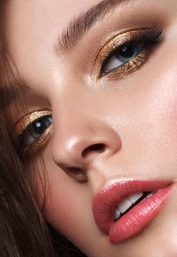 Primo piano di un volto con ombretto bronzo, ciglia definite, pelle liscia e labbra rosa lucide. Presenta riflessi sottili e sopracciglia naturali.