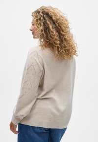 Beige knit sweater with a loose fit, featuring textured diamond patterns on the sleeves and ribbed cuffs, paired with blue denim jeans.
