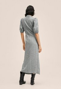 Femme portant une robe midi grise avec des manches courtes bouffantes et des bottes noires mi-mollet, se tenant de dos contre un fond uni.