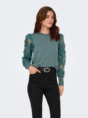 Femeie purtând un top verde-albăstrui cu mâneci lungi, transparente și volănașe, și pantaloni negri cu talie înaltă şi curea neagră, stând cu o mână în buzunar.