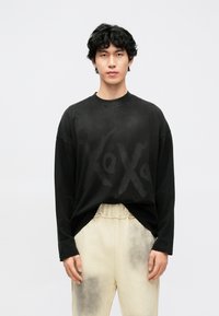 Melna garām piedurknēm T-krekls ar izbalējušu teksta apdruku. Audums ir mīksts, un piegriezums ir brīvs, kombinēts ar gaišā bēša krāsas biksēm ar tumšiem plankumiem.