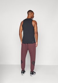 Zwarte mouwloze tanktop gecombineerd met bordeauxrode sweatpants met zwarte diagonale strepen en zwarte sportschoenen. Neutrale achtergrond.