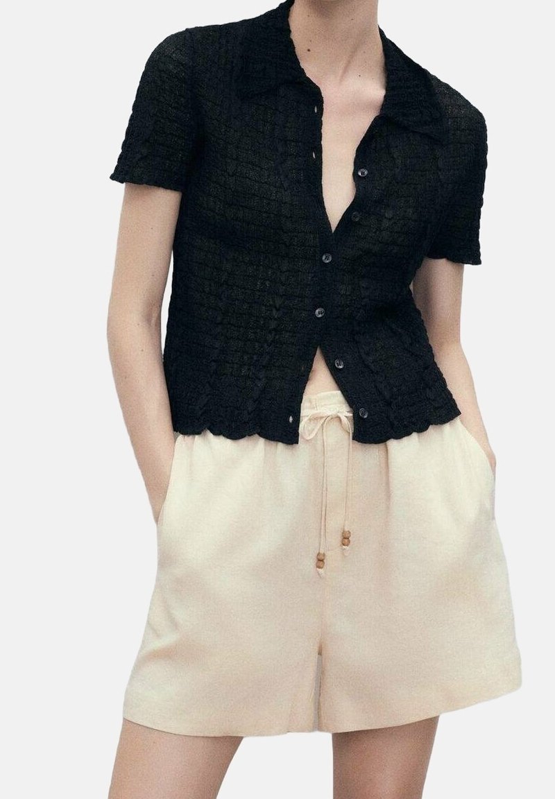 Femme portant une chemise noire texturée à manches courtes boutonnée et un short beige clair à cordon, les mains dans les poches.