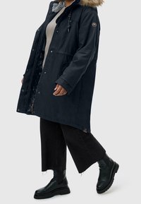 Person som bär en marinblå parkas med päls huva, beige topp, korta svarta vida byxor och svarta kraftiga läderstövlar som går mot höger.