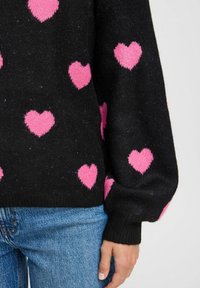 Maglione nero con motivi a cuore rosa pelosi, con orlo e maniche a coste. Indossato sopra jeans in denim blu.