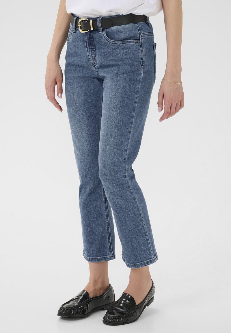 Femme portant un jean bleu droit avec une ceinture noire et des mocassins en cuir noir, debout sur un fond blanc.