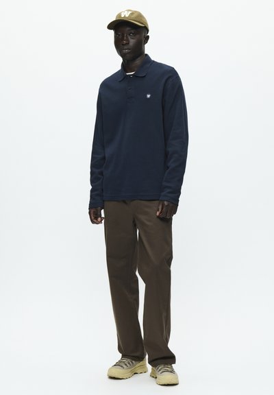Polo shirt a maniche lunghe blu scuro con un piccolo logo, abbinato a pantaloni marroni e un cappellino beige con un'emblema bianco. Sneakers chiare.