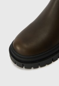 Botín de cuero marrón hasta el tobillo con punta redondeada, parte superior texturizada y una suela gruesa de goma negra con surcos profundos para mayor tracción.