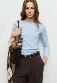 Giovane donna in maglione azzurro chiaro e pantaloni marroni, che tiene una sciarpa a quadri marrone e una borsa a tracolla su uno sfondo neutro.