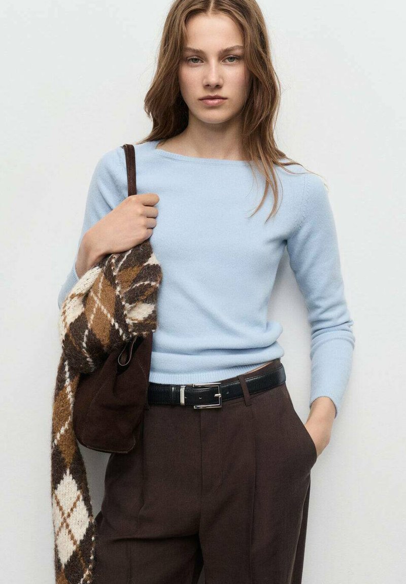 Giovane donna in maglione azzurro chiaro e pantaloni marroni, che tiene una sciarpa a quadri marrone e una borsa a tracolla su uno sfondo neutro.