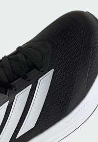 Nahaufnahme eines schwarzen Sneakers mit weißen Streifen, schwarzen Schnürsenkeln und einer strukturierten Sohle mit der Aufschrift „Cloudfoam“ an der Seite.