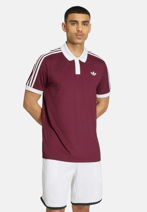 Ein junger Mann steht mit den Händen hinter dem Rücken, trägt ein bordeauxrotes Poloshirt mit weißem Kragen und Streifen, kombiniert mit weißen Shorts mit schwarzem Besatz.