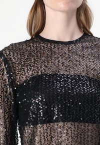 Schwarzes Paillettenoberteil mit einem transparenten Überzug und einem durchgehenden schwarzen Paneel über der Brust. Hat einen runden Ausschnitt und eine strukturierte, glitzernde Oberfläche.