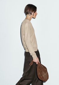 Beige Pullover mit Rundhalsausschnitt und langen Ärmeln, kombiniert mit weiter geschnittenen, dunkelbraunen Hosen. Hält eine strukturierte, braune Perlenhandtasche.