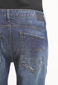 Vaqueros de mezclilla en un lavado azul oscuro con efectos visibles de desgaste. Presentan dos bolsillos traseros con detalles de costura y una etiqueta de marca en la pretina.