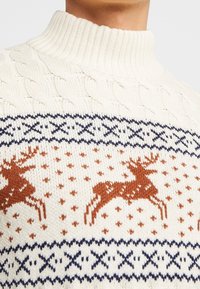 Krämfärgad stickad tröja med hög krage, kabelmönster och en festlig design med bruna renar och marinblå detaljer.