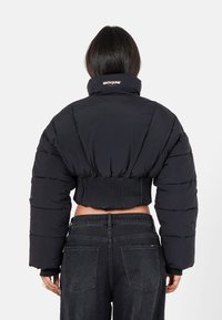 Femme portant une veste en duvet noire courte et un jean foncé taille haute, vue de dos sur un fond uni.