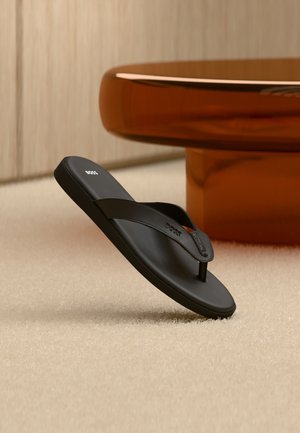 Schwarze BOSS Flip-Flop Sandale auf einem beigen Teppich mit einem großen runden bernsteinfarbenen Tisch im Hintergrund.