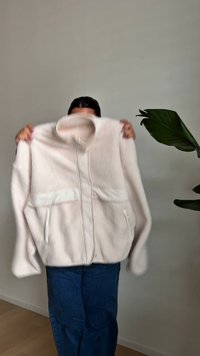 Osoba drží béžovú fleecovú bundu so stojatým golierom a vreckami na zips na pozadí obyčajnej sivej steny a drevenej podlahy.