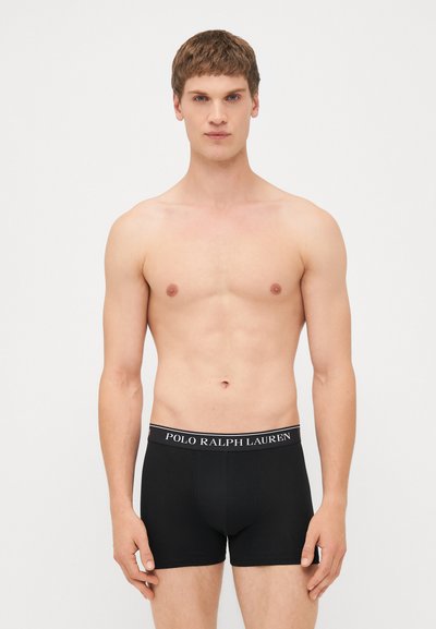 Črne bokserice iz mehkega blaga z širokim elastičnim pasom, na katerem piše "POLO RALPH LAUREN." Model ima nevtralen izraz.